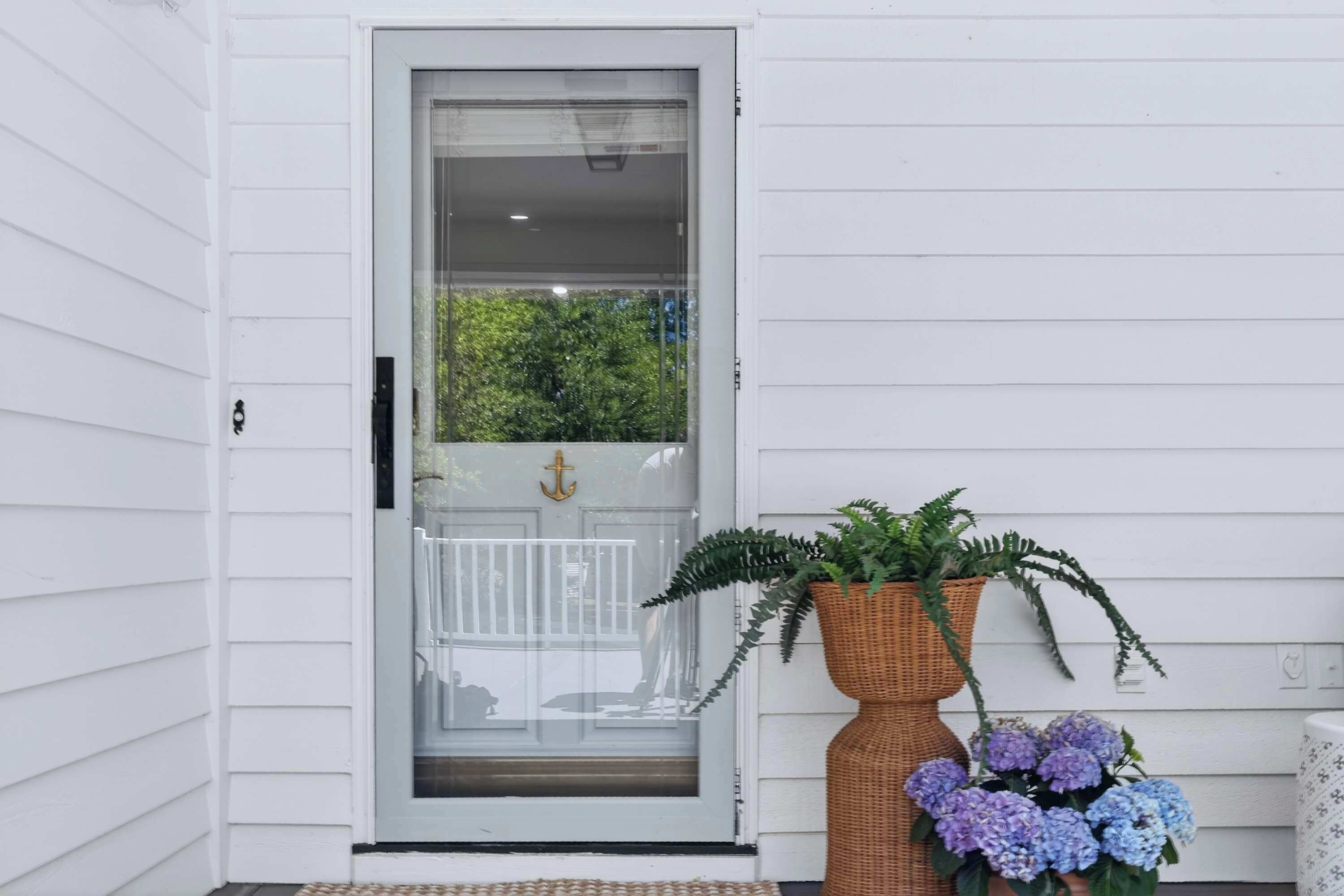 A white door with a glass panel, a wicker planter with greenery, and purple flowers on the porch.
