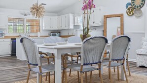 Un coin repas avec une table et des chaises blanches, une cuisine en arrière-plan et un miroir au mur.