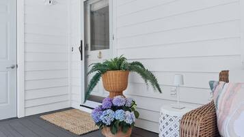 A white exterior wall, a door, a window, a potted plant, a small table with a lamp, and a wicker chair.