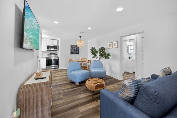 Una sala de estar moderna con un televisor de pantalla plana, sofás azules, una mesa de centro de mimbre y una cocina al fondo.