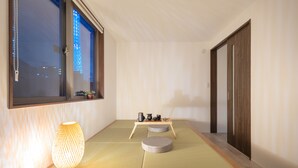 Une pièce au sol recouvert de tatamis, une petite table avec une lampe et une fenêtre donnant sur le ciel nocturne.