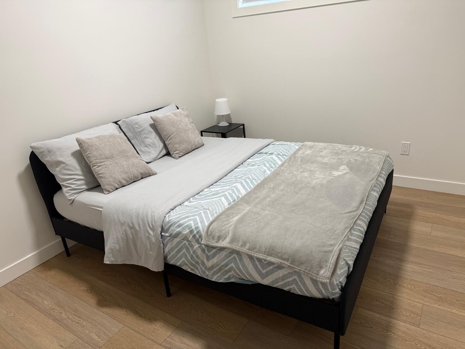 Una cama bien tendida con un edredón estampado, una mesita de noche con lámpara y una ventana con persianas.