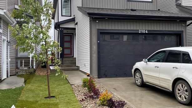 Een tweelaags huis met een garage, oprit en een gazon met een kleine boom.