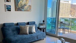 Een moderne woonkamer met een blauwe bank, een glazen salontafel en een balkon met uitzicht op gebouwen.