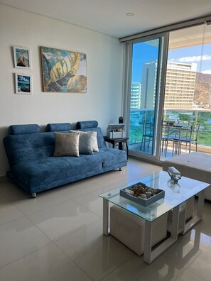 Ein modernes Wohnzimmer mit einem blauen Sofa, einem Glastisch und einem Balkon mit Blick auf Gebäude.