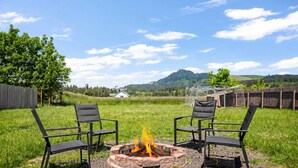 Feuerstelle mit zwei Stühlen, umgeben von einer grünen Wiese, mit Holzzaun und einem Haus im Hintergrund.