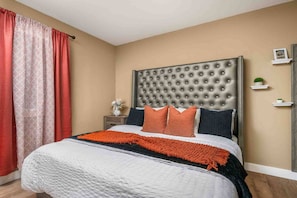 Ein Schlafzimmer mit einer gepolsterten Kopfteile, einem Bett mit weißer und orangefarbener Decke, einem Nachttisch mit einer Vase und einem kleinen Regal mit einer Pflanze.