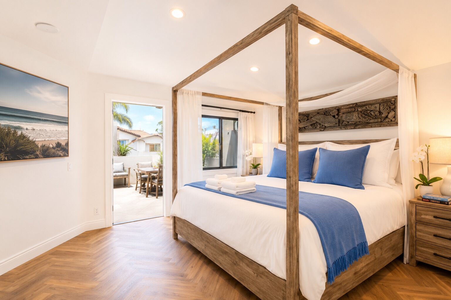 Una cama con dosel de madera, ropa de cama blanca y almohadas azules. Hay una mesita de noche de madera con una lámpara y una pequeña planta. El cuarto tiene piso de madera y vista al patio exterior.