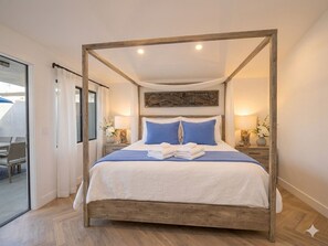 Ein Bett mit Baldachin, ausgestattet mit weißer Bettwäsche und blauen Kissen, zwei Nachttischlampen und Blick auf den Außenbereich durch eine Glastür.