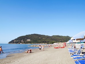 Ein Strand mit Liegestühlen und Sonnenschirmen, eine felsige Küstenlinie und eine Hügelkulisse im Hintergrund.