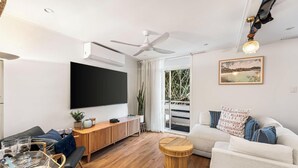 Ein Wohnzimmer mit Flachbildfernseher, Deckenventilator und großem Fenster mit Blick auf Grünflächen.