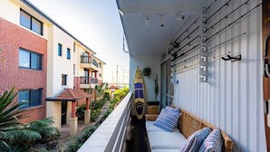 Ein schmaler Balkon mit einer Couch und einem Surfbrett, einer Brüstung sowie ein Gebäude mit zahlreichen Fenstern und Balkonen.