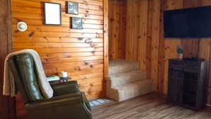 Ein gemütliches Blockhaus-Interieur mit Holzwänden, einem Ledersessel, einem kleinen Tisch mit Tasse, einer Treppe und einem Fernseher auf einem Holzschrank.