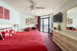 Ein Hotelzimmer mit einem großen Bett, einem Schreibtisch, einem Sessel, einem Fernseher, einem Deckenventilator und einem Balkon mit Blick.