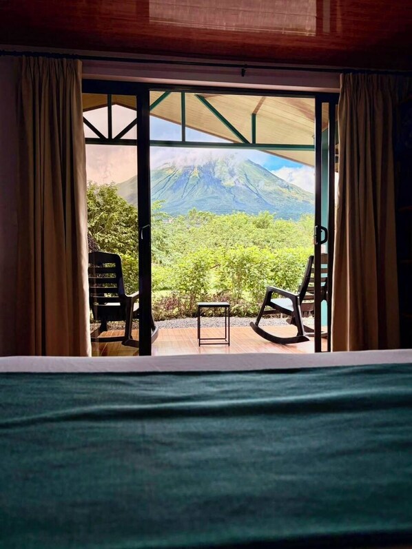 A room with a bed, two chairs, and a table. There is a large window with curtains, through which a mountainous landscape is visible.