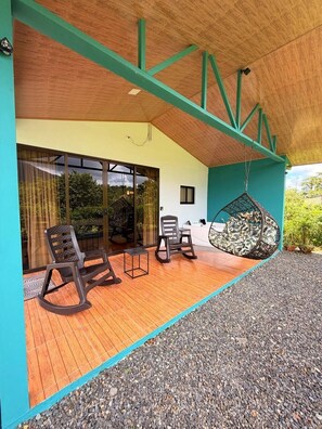 Un espacio de estar al aire libre techado, con piso de madera, sillas, una hamaca colgante y vistas de la vegetación circundante.