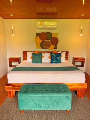 Habitación de hotel con cama, un banco tapizado en verde, cabecero de madera, dos mesitas de noche y un equipo de aire acondicionado de pared.