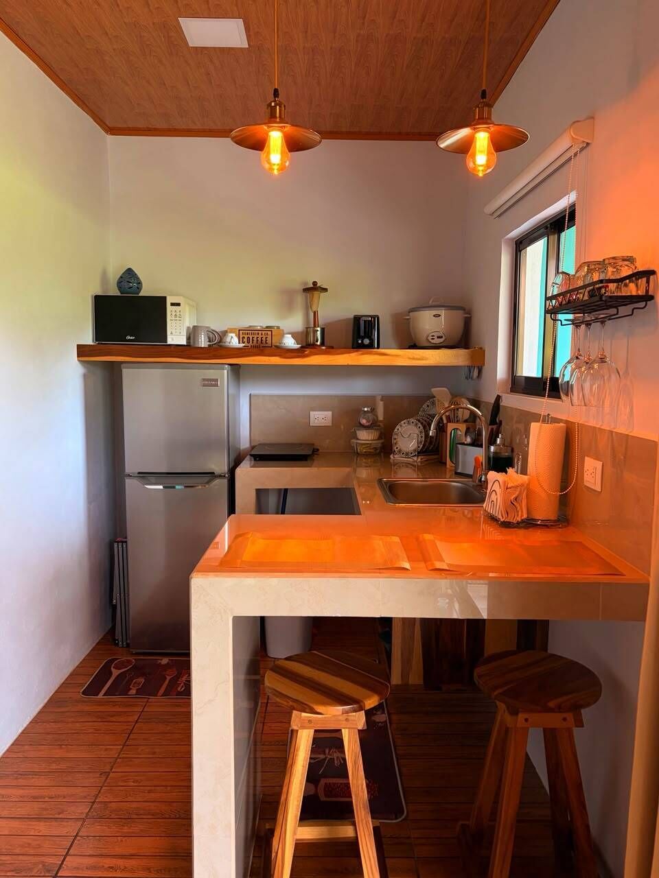 Una cocina compacta con encimera de madera, dos taburetes, un refrigerador y luces colgantes.