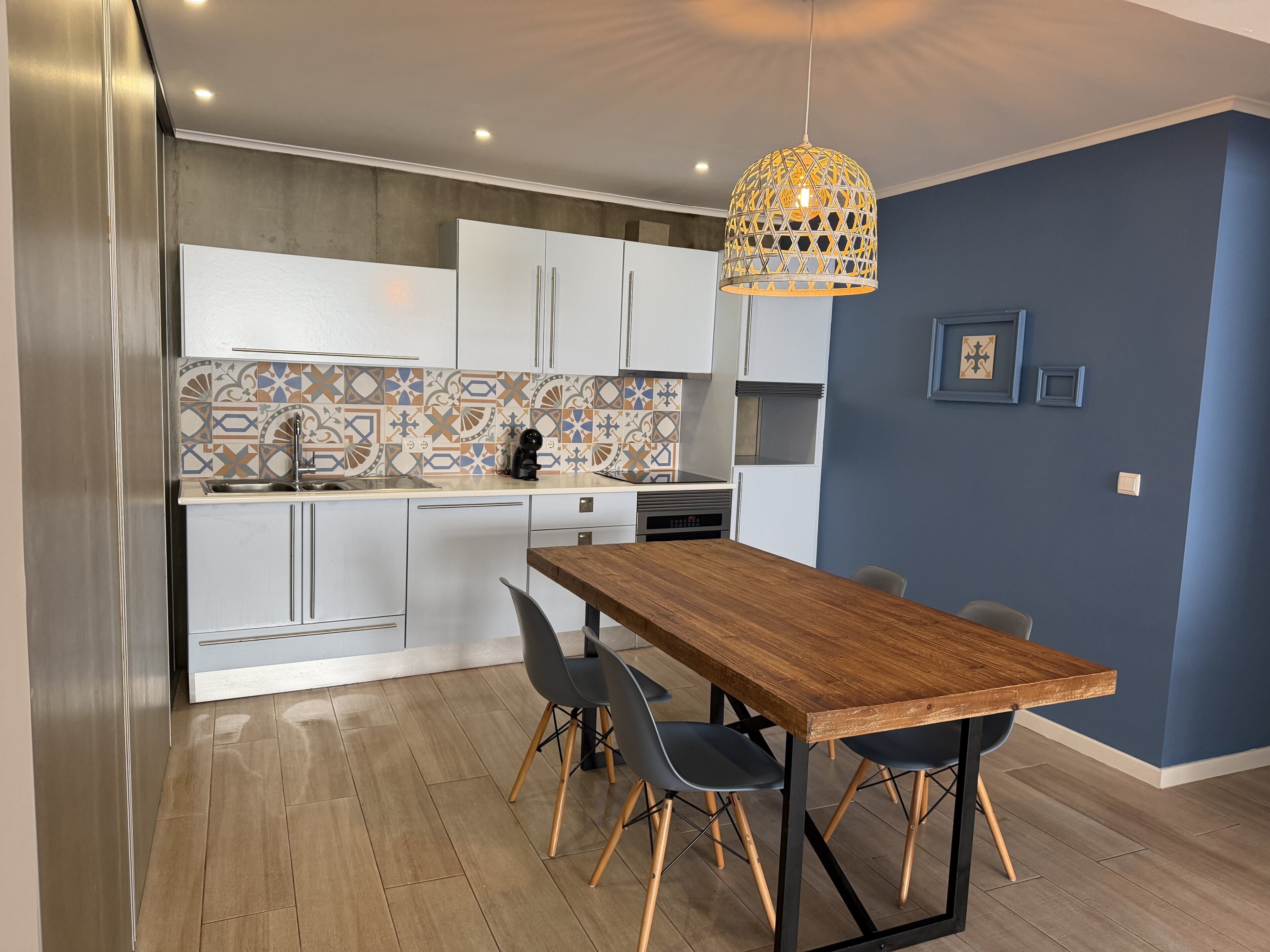 Een moderne keuken met witte kastjes, een backsplash met patroon, een houten eettafel met stoelen en een hangende hanglamp.