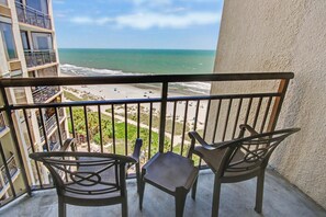 A balcony with two chairs and a small table, overlooking a beach with a view of the ocean and a building in the background. - Oceanfront King Studio + Beach Access + Lazy River (Myrtle Beach)