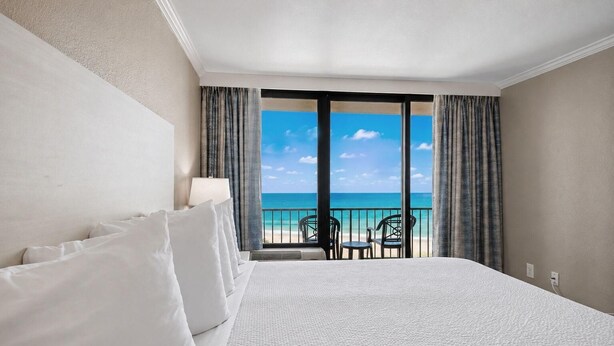 A hotel room with a large bed, a view of the beach, and a balcony with chairs. - Oceanfront King Studio + Beach Access + Lazy River (Myrtle Beach)