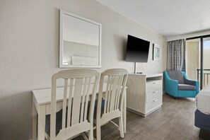 A hotel room with a desk, two chairs, a television, a bed, and a window with curtains. - Oceanfront King Studio w/ Sleeper Sofa + Pools (Myrtle Beach)