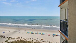 Vista de una playa desde un balcón, con cielo despejado, arena y sombrillas.