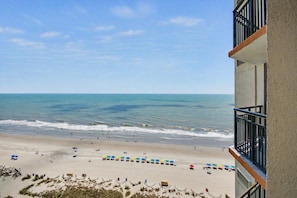 Blick auf einen Strand vom Balkon, mit blauem Himmel, Sand und Sonnenschirmen.
