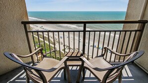 A balcony with two chairs and a table, overlooking a beach with people and vehicles. - Oceanfront King Studio w/ Sleeper Sofa + Pools (Myrtle Beach)