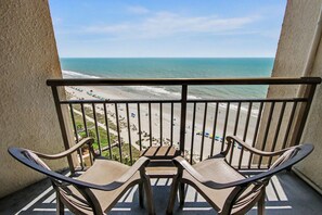 A balcony with two chairs and a table, overlooking a beach with people and vehicles. - Oceanfront King w/Balcony & Kitchen | Dog Friendly (Myrtle Beach)