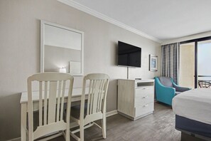 A hotel room with a desk, two chairs, a television, a bed, a window with curtains, and a small cabinet. - Oceanfront King Studio w/ Sleeper Sofa + Pools (Myrtle Beach)