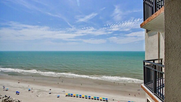 A beach view from a balcony with a clear sky, ocean, and beach umbrellas. - Oceanfront King Studio w/ Sleeper Sofa + Pools (Myrtle Beach)