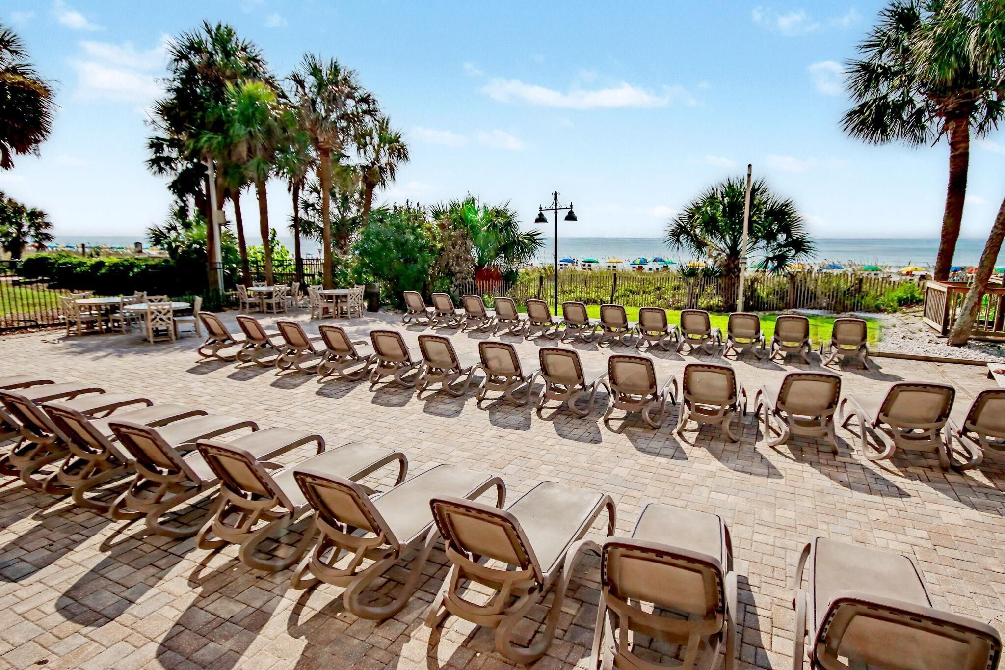A beachside lounge area with rows of sun loungers and tables, palm trees, and a view of the ocean.