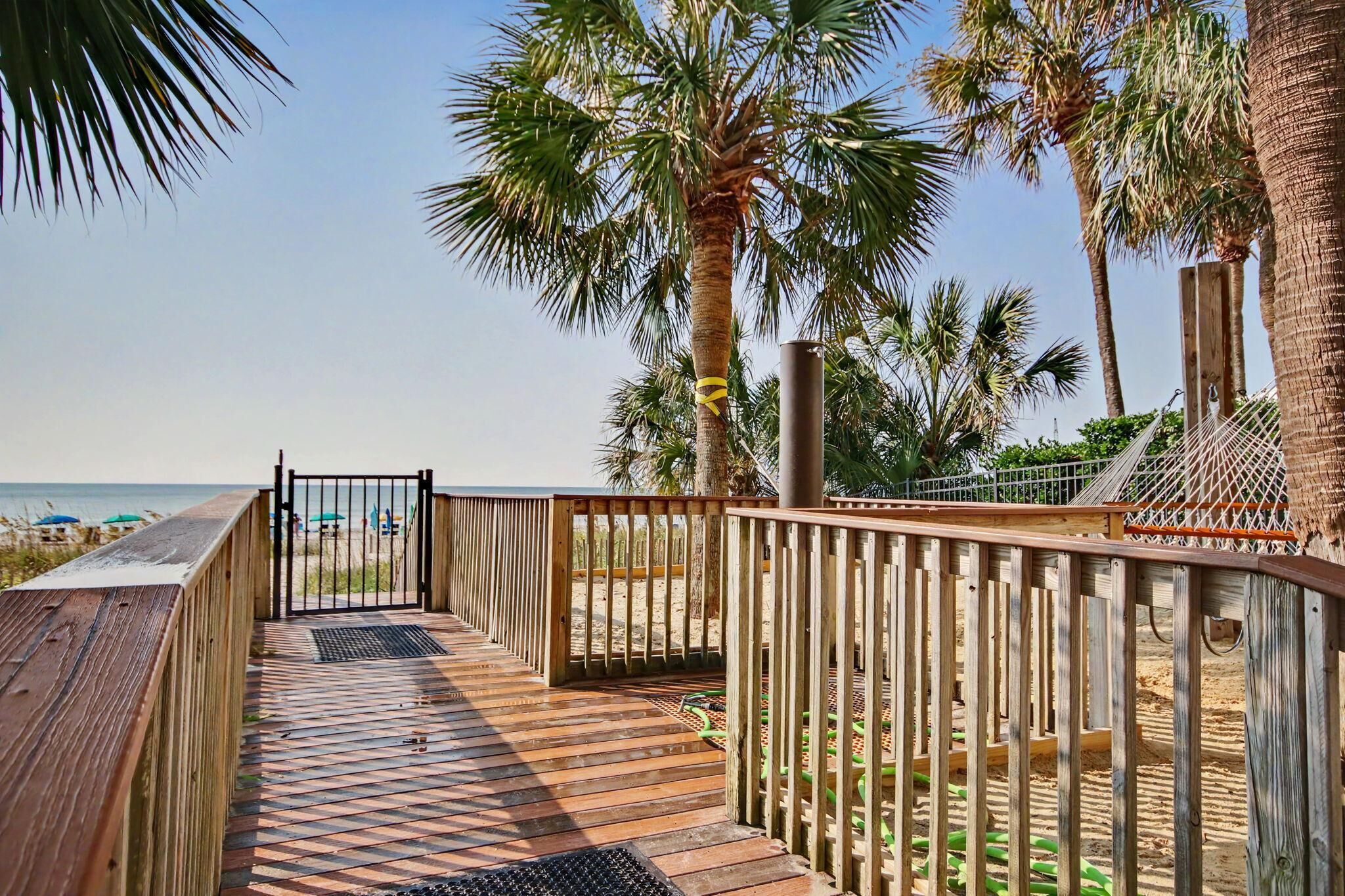 A wooden deck leading to a beach with palm trees and a clear sky.