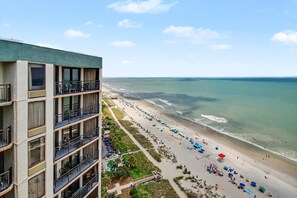 A high-rise hotel with balconies overlooking a beach with umbrellas and people. - Oceanfront King Studio w/ Sleeper Sofa + Pools (Myrtle Beach)