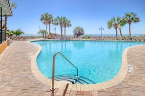 A pool with clear blue water, surrounded by lounge chairs and palm trees, with a view of the ocean. - Oceanfront King Studio w/ Sleeper Sofa + Pools (Myrtle Beach)