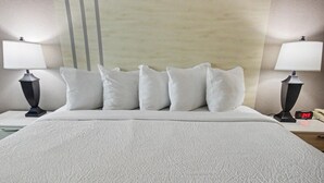 Habitación de hotel con cama, ropa de cama blanca, dos lámparas de noche, una mesita de noche con un reloj y una pared con líneas verticales.
