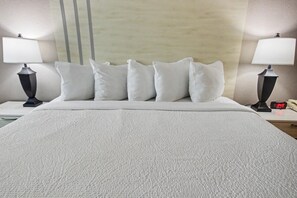 A hotel room with a bed, white bedding, two bedside lamps, a nightstand with a clock, and a wall with vertical lines. - Oceanfront King Studio w/ Sleeper Sofa + Pools (Myrtle Beach)