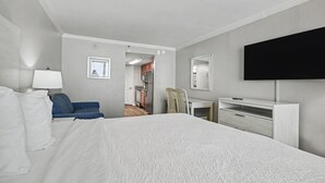 A hotel room with a bed, a desk, a chair, a TV, and a door leading to a kitchen area. - Oceanfront King Studio w/ Sleeper Sofa + Pools (Myrtle Beach)