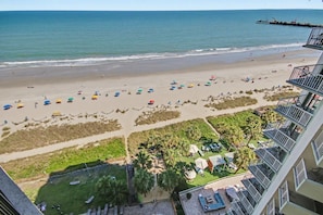 A beach view from a high vantage point, including a hotel with balconies, a sandy beach with umbrellas, and a pier in the distance. - Oceanfront Studio w/ Two Queens + Beach & Pools (Myrtle Beach)