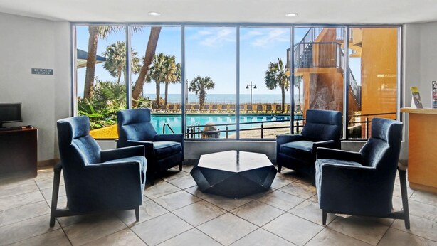 Una hall d'albergo moderna con vista piscina, due poltrone blu, un tavolino esagonale e un cartello per il business center.