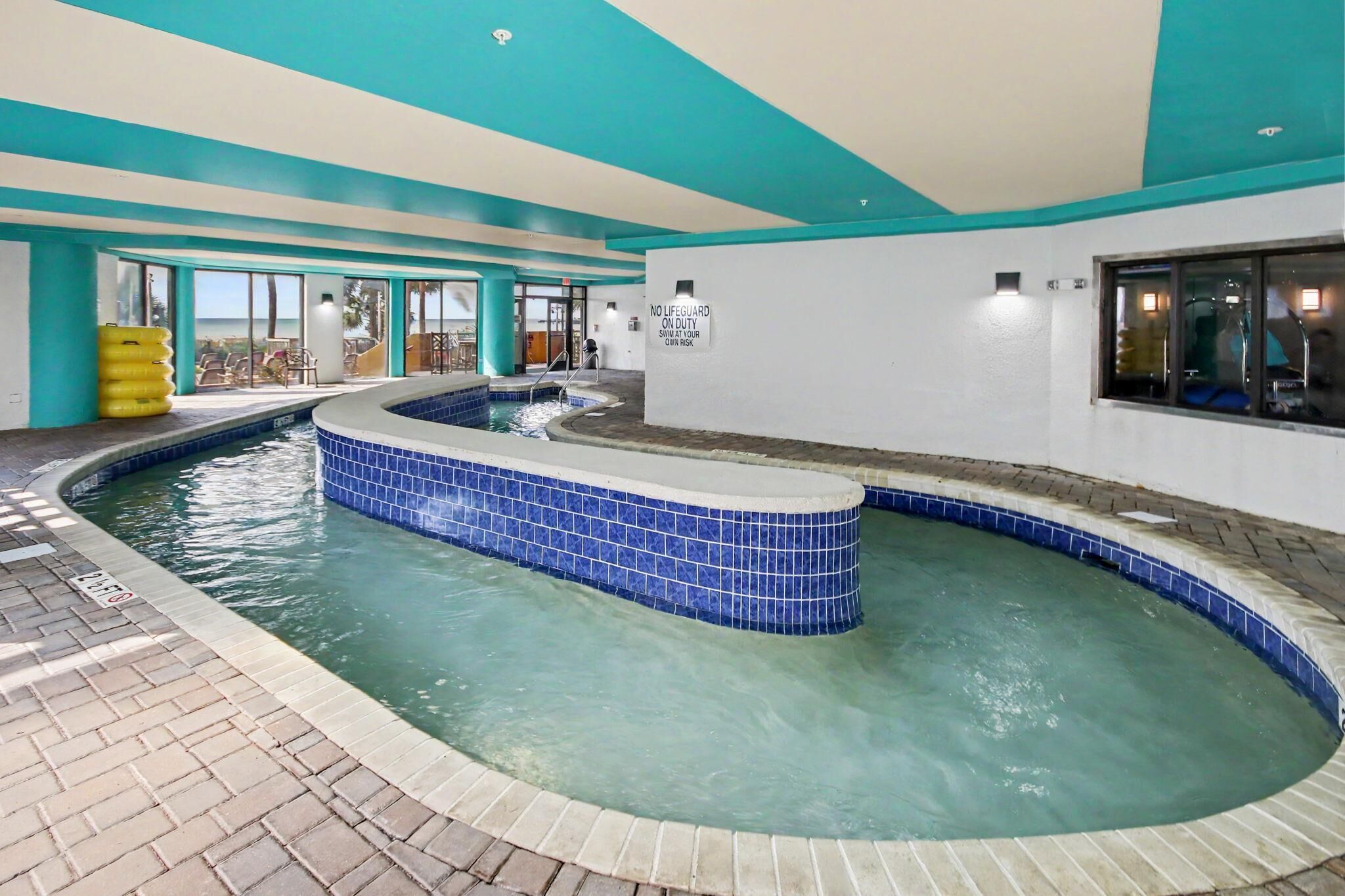 En indendørs swimmingpool med et bølget design, et skilt for livredder og udsigt til det fri.