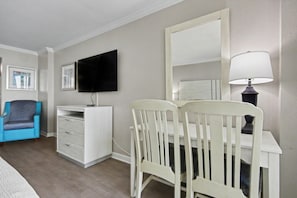 A room with a white desk, a television mounted on the wall, a blue chair, and a lamp. - Oceanfront Studio w/ Two Queens + Beach & Pools (Myrtle Beach)