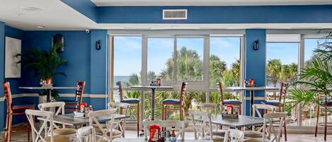 A dining area with tables and chairs, large windows, and a view of palm trees.