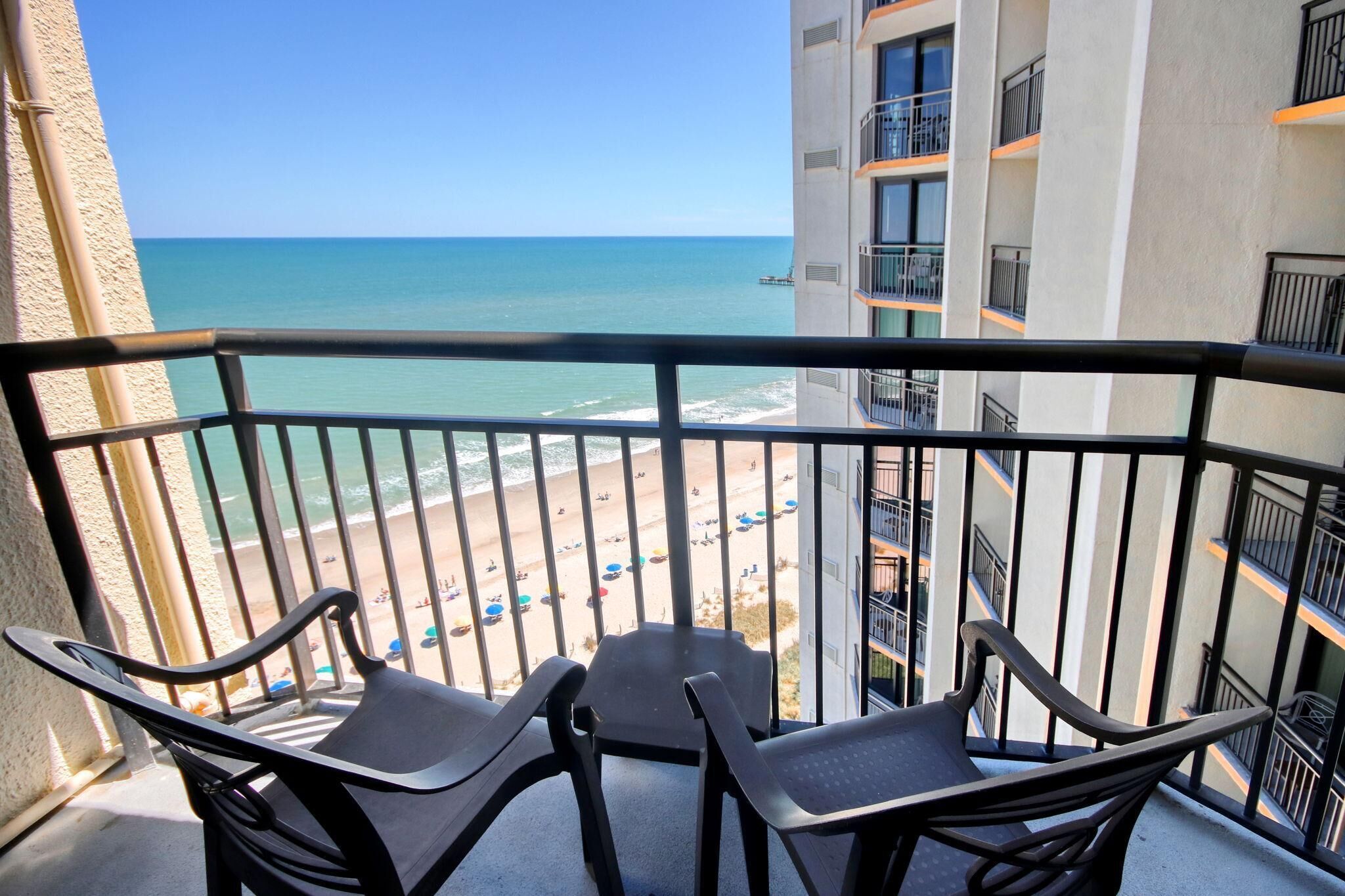 A balcony with a view of a beach and ocean. There are two chairs and a table on the balcony.