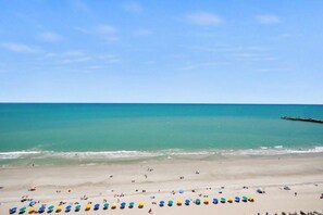A beach with a pier extending into the water, people relaxing on the sand, and umbrellas lined up along the shore. - Oceanfront Studio w/ Two Queens + Beach & Pools (Myrtle Beach)