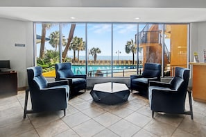 A modern hotel lobby with a pool view, two blue armchairs, a hexagonal coffee table, and a business center sign.