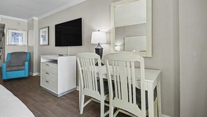 A hotel room with a bed, a desk with two chairs, a TV, a lamp, and a small cabinet. - Oceanfront Studio w/ Two Queens + Beach & Pools (Myrtle Beach)