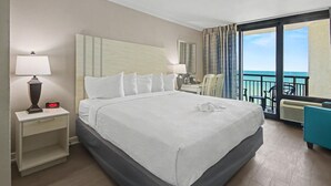 A hotel room with a bed, bedside tables, a lamp, a clock, a chair, a table, and a view of the ocean. - Ocean View King Room + Beach Access + Lazy River (Myrtle Beach)