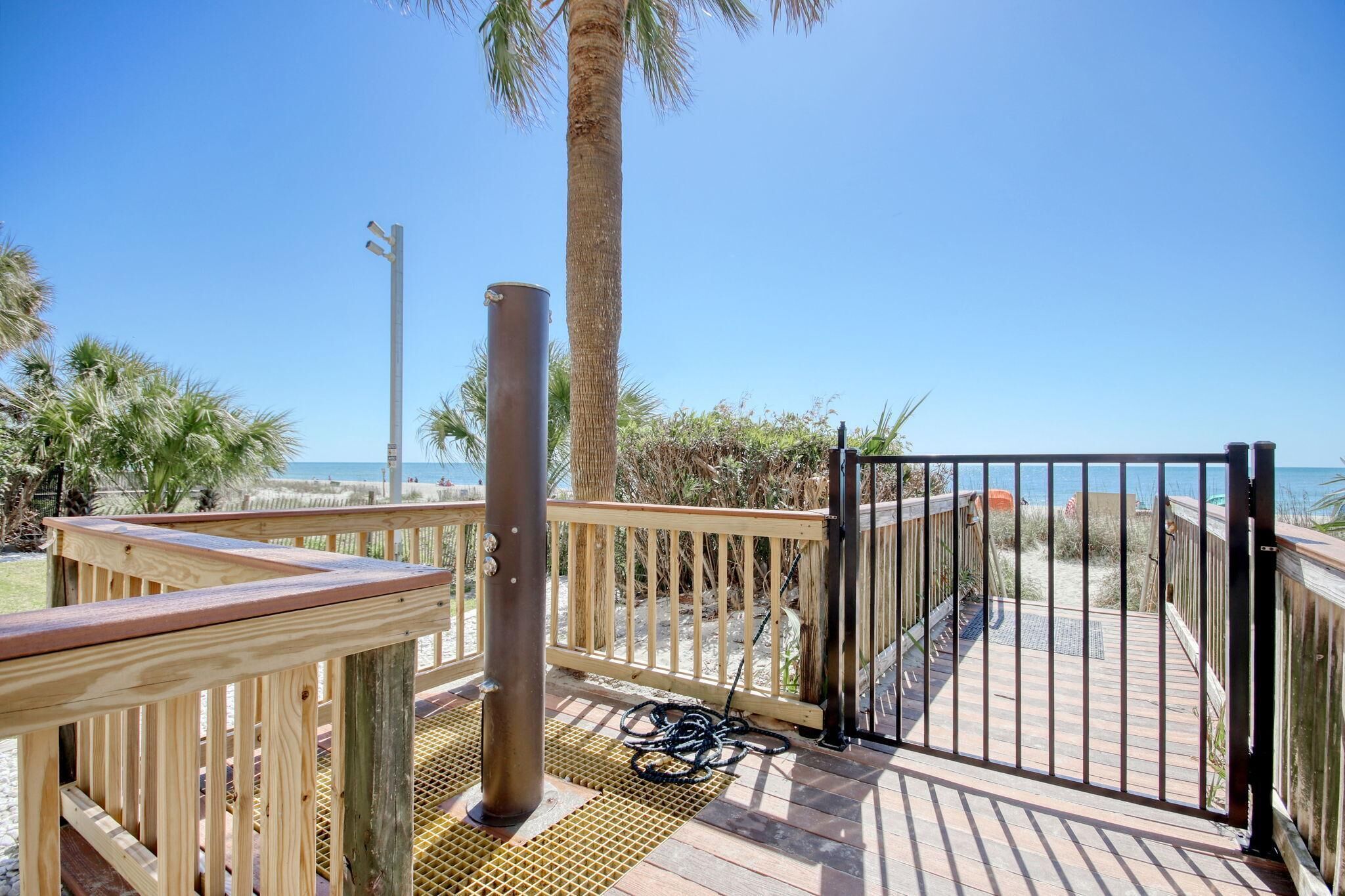 A wooden deck with a metal gate leading to a beach, palm trees, and a clear sky.